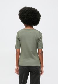 Άτομο με σκούρα σγουρά μαλλιά που φοράει πράσινο κοντομάνικο πουκάμισο και μαύρα τζιν, στέκεται με το πρόσωπο στραμμένο μακριά σε έναν απλό ανοιχτό φόντο.