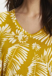 Keltainen lyhythihainen paita, jossa on V-pääntie ja valkoinen lehtikuosi. Kangas näyttää pehmeältä ja sileältä.