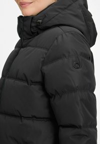 Nahaufnahme einer Person in einer schwarzen Steppjacke mit Kapuze, Reißverschluss vorne und einem kreisförmigen Logo auf dem oberen Ärmel.
