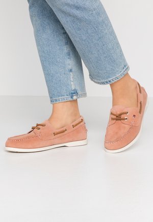 Persoon die een lichtblauwe spijkerbroek draagt en roze suède mocassins in mocassinstijl met bruine veters en witte zolen, staand op een witte vloer.