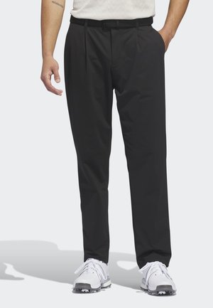 Homme portant un pantalon noir droit avec passants de ceinture et des chaussures de golf blanches, debout les mains dans les poches sur un fond uni.