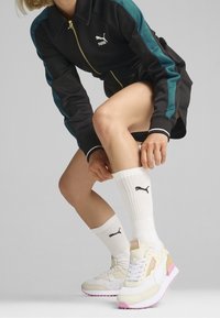 Puma sneakers in beige and pink with textured upper. Paired with white socks featuring a logo, and a black zip-up jacket with teal accents.