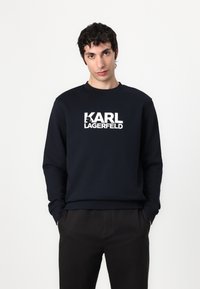 Navy plavi dukserica s okruglim izrezom i dugim rukavima, s istaknutim bijelim logotipom "Karl Lagerfeld" na prednjem dijelu.