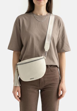 Woman wearing brown t-shirt and pants, carrying off-white crossbody bag with silver zipper and tassel, wide branded strap over shoulder.