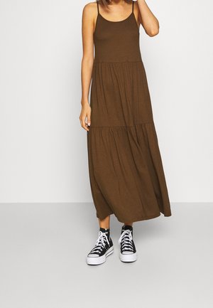 Femme portant une robe maxi marron à volants sans manches et des baskets montantes noires avec des lacets blancs, debout devant un fond uni.