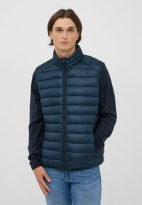 Gilet di piuma navy con collo alto, caratterizzato da un design trapuntato e chiusura a zip, abbinato a maniche lunghe scure e jeans azzurri chiari.