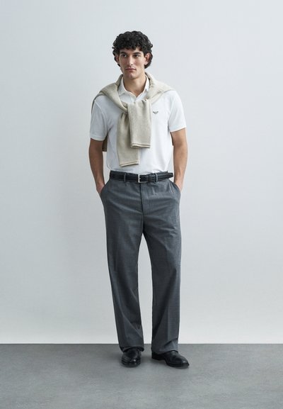 Biele polo tričko s logom, sivé nohavice a béžový pletený sveter zviazaný cez ramena. Čierny opasok a topánky dotvárajú vzhľad.