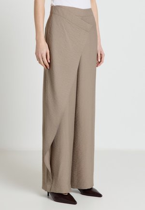 Pantaloni ampi color taupe con tessuto strutturato e pannello frontale sovrapposto, indossati con tacchi scuri a punta e top bianco infilato.