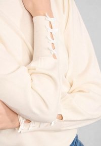 Maniche di un maglione color crema con tagli verticali legati con piccoli fiocchi bianchi, indossato con le braccia incrociate sul petto.