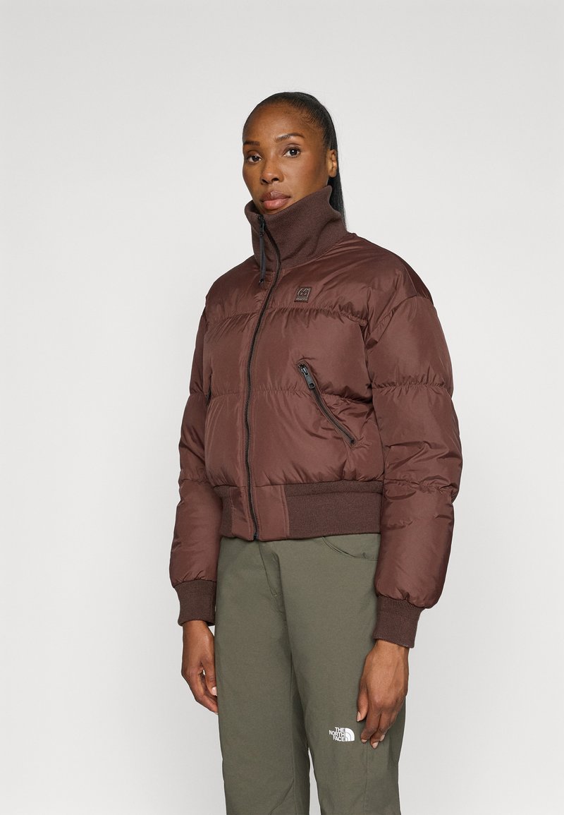 Casaco puffer castanho com gola alta, fecho de correr e bolsos laterais. Punhos e bainha canelados. Modelo a usar calças verdes-oliva. Fundo simples.