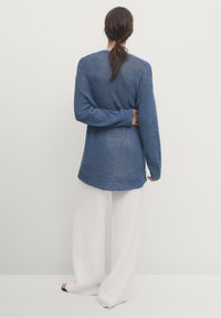 Person med brunt hår som är uppsatt, iklädd en blå stickad tröja och lösa vita byxor, stående barfota mot en enkel vit vägg.