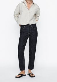 Vaaleanharmaa nappimyynti-paita, jossa on käärityt hihat, tummansiniset farkut, joissa on kontrastiväriommeltu sauma, ja mustat flip-flopit tasaisella taustalla.