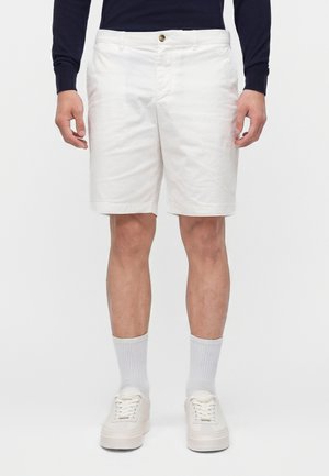 Homme portant un short blanc jusqu’aux genoux, des baskets blanches, des chaussettes blanches mi-hautes et un haut à manches longues sombre, debout devant un fond uni.