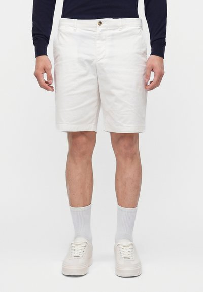 Homme portant un short blanc jusqu’aux genoux, des baskets blanches, des chaussettes blanches mi-hautes et un haut à manches longues sombre, debout devant un fond uni.