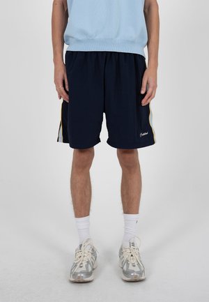 Personne portant un short de sport bleu marine avec des bandes latérales bleu clair et jaune, des chaussettes blanches et des chaussures de course grises, debout devant un fond uni.