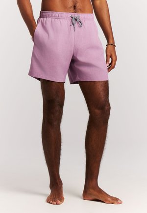 Hombre con pantalones cortos de baño de color lila claro con cordón blanco y negro, de pie descalzo sobre un fondo liso.