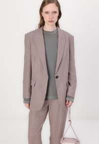 Rutete rosa og grå blazer med en enkelt knapp, kombinert med en grå hettegenser. Modellen holder en liten rosa veske. Jevn tekstur.