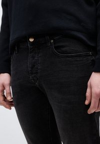 Κοντινό πλάνο ενός ατόμου που φοράει μαύρο τζιν και μαύρο μακρυμάνικο τοπ, με τα χέρια να είναι χαλαρά στο πλάι.