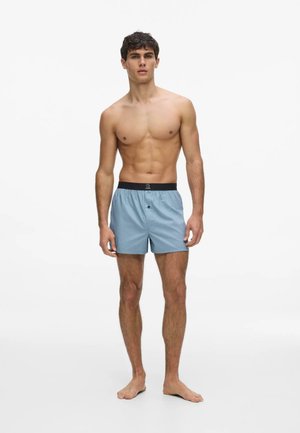 Jeune homme debout pieds nus portant un boxer bleu clair avec une ceinture élastique noire sur fond blanc.