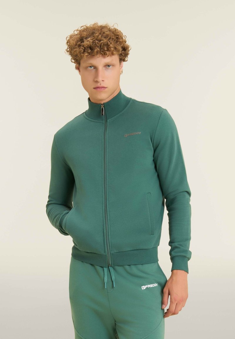 Felpa verde con zip e colletto alto, polsini a costine e tasche laterali. Presenta un dettaglio con logo sul petto e un tessuto morbido e texturizzato.