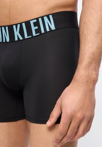 Κοντινό πλάνο ενός άνδρα που φοράει μαύρα εσώρουχα μπόξερ Calvin Klein με ελαφρώς γαλάζια λογότυπο στη μέση, δείχνοντας το σώμα και το χέρι να ακουμπάει στο μηρό.