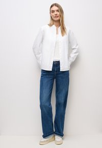 Camisa blanca de botones con cuello tipo camisa, combinada con jeans rectos azules. Modelo lleva zapatillas blancas y está de pie contra un fondo neutro.