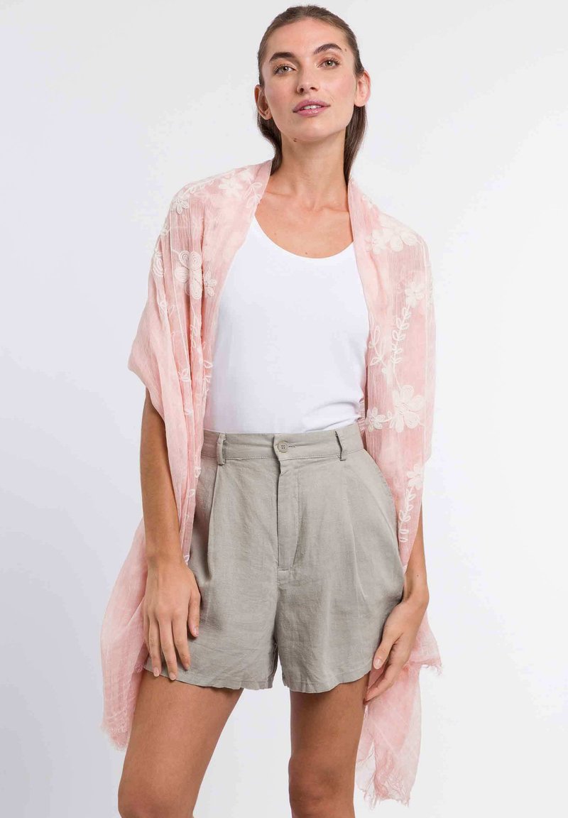 Șal de un roz deschis cu broderie florală, fabricat dintr-un material ușor, cu un design deschis în față, marginile franjurate, asortat cu un top alb și pantaloni scurți bej.