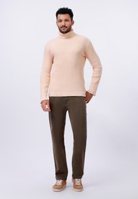 Gebreide coltrui in licht perzik, gecombineerd met olijfgroene broek. Opvallende textuur en ribbeldetails op de mouwen. Bruine sneakers.