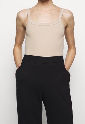 Osoba nosząca beżowy prążkowany tank top włożony do czarnych spodni z wysokim stanem, z rękami w kieszeniach, stojąca na jednolitym tle.