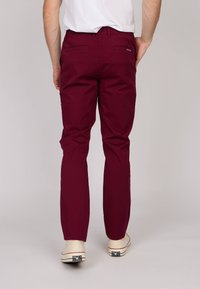 Pantalons en coton bordeaux avec une coupe slim, deux poches arrière et une texture lisse. Associés à des chaussures blanches avec des accents rouges.