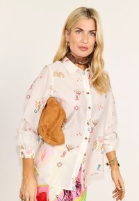 Ξανθιά γυναίκα με χαλαρά κυματιστά μαλλιά φοράει λευκό πουκάμισο με πολύχρωμα σχέδια εμπνευσμένα από τη δυτική θεματολογία και κρατάει μικρό τσαντάκι από ταμπά σουέντ.