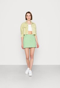 Saia mini em padrão verde ondulado feita de tecido suave, combinada com uma jaqueta curta de corduroy verde-claro e uma t-shirt cropped branca ajustada. Ténis brancos.