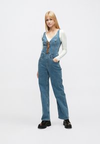 Ολόσωμη φόρμα από τζιν σε ανοιχτό μπλε χρώμα, με βαθύ ντεκολτέ, ρυθμιζόμενα κορδόνια και ορθογώνιες τσέπες· φορεμένη πάνω από λευκό μπλουζάκι με μακριά μανίκια.