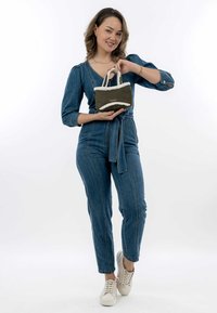 Oliven grøn håndtaske med hvid, fluffy kant og dobbelt håndtag; model iført en denim jumpsuit med lige ben og lyse sneakers.
