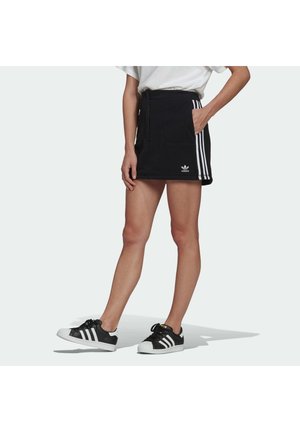 Osoba nosząca czarną mini spódniczkę Adidas z białymi paskami oraz czarne buty sportowe Adidas z białymi paskami na jasnym tle.