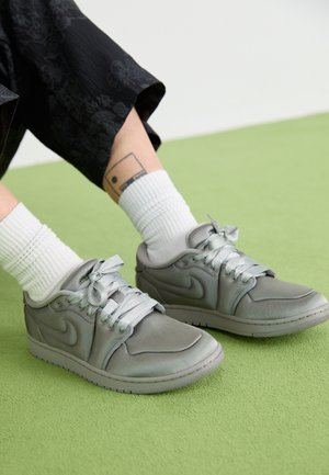 Persona con calcetines blancos y zapatillas Nike grises sobre una alfombra verde, mostrando un tatuaje en el tobillo y pantalones negros con estampado floral.