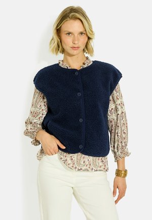 Mujer rubia que lleva un chaleco polar azul marino con botones sobre una blusa estampada, pantalones blancos y pulseras doradas en su muñeca derecha.
