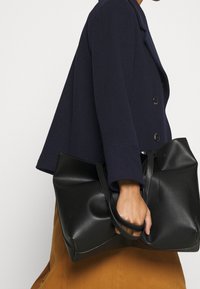 Sac en toile en cuir noir avec deux poignées robustes et une surface texturée, tenu d'une main à côté d'une veste marine et d'une jupe marron.