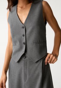 Veste en tissu gris avec un col en V, cinq boutons noirs et deux poches. Texture lisse avec une coupe ajustée et un pantalon gris assorti.