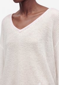 Personne à la peau foncée portant un pull rose clair, tricoté de façon lâche, avec un col en V, montrant le cou et le haut de la poitrine.