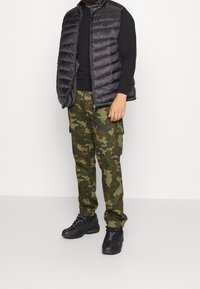 Svart puffy väst över en svart långärmad tröja, parat med gröna kamouflagemönstrade cargobyxor och svarta sneakers. Har dragkedjestängning och fickor.