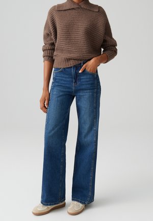 Osoba w brązowym, prążkowanym swetrze, wysokich, szerokich, niebieskich dżinsach i białych trampkach, z jedną ręką w kieszeni na szarym tle.