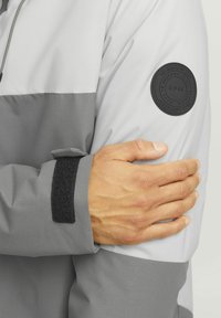 Hallgray ja heledamat hall jakki tekstuurse kangaga. Õlal on ümmargune must pidin ja mansetil on Velcro riba.