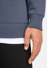 Sudadera azul con puños y dobladillo acanalados, superpuesta sobre una camisa blanca, combinada con pantalones negros. Enfocándose en una tela elegante y transpirable.