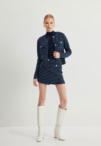 Marineblaues Strickjackett und passender Rock mit weißen Streifen. Die Jacke hat Knöpfe, und das Modell trägt weiße knielange Stiefel.