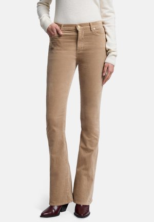 Jeans in velluto a coste beige a gamba svasata con zip frontale, tasche anteriori e una leggera lucentezza. Abbinati a un top chiaro e stivali neri alla caviglia.