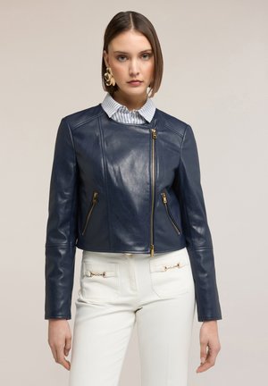 Frau trägt eine marineblaue Lederjacke mit asymmetrischen goldenen Reißverschlüssen, weiße hoch taillierte Hosen mit goldenen Details und große goldene Ohrringe.