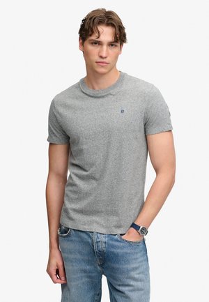 Giovane uomo che indossa una maglietta grigio chiaro a maniche corte, jeans blu e un orologio da polso blu scuro, con una mano in tasca.