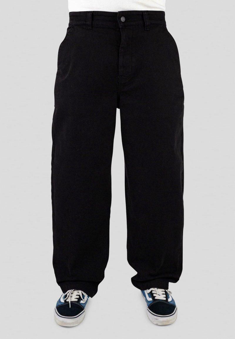 Vue de face d'une personne portant un pantalon noir ample et des baskets blanches bleu-noir à lacets sur un fond uni.
