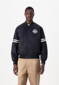 Chaqueta bomber en color navy con un logo en el pecho, detalles en blanco en los puños y cierre de botones, fabricada con un tejido liso y duradero.